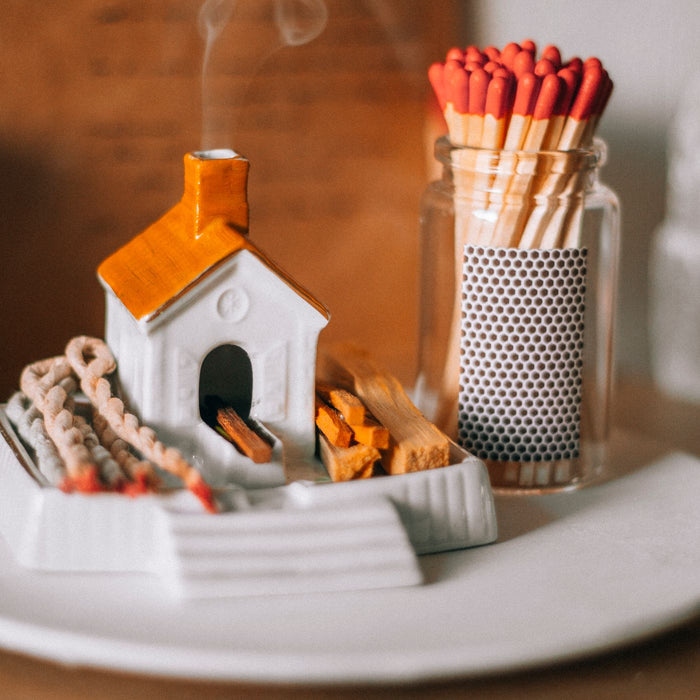A ceramic incense burner shaped like a small house, with smoke coming from it and matches lying beside it.