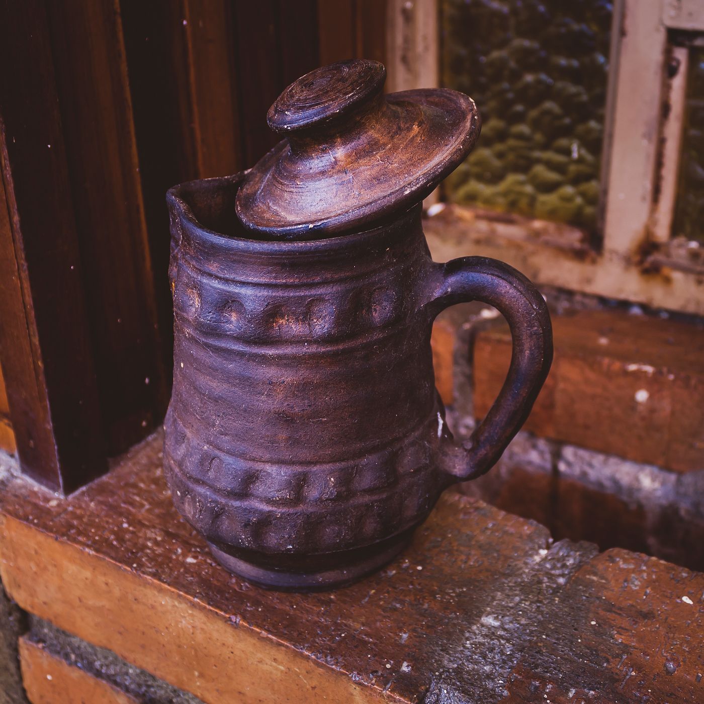 A rustic antique style jug resting on a brick ledge near a wooden wall.
