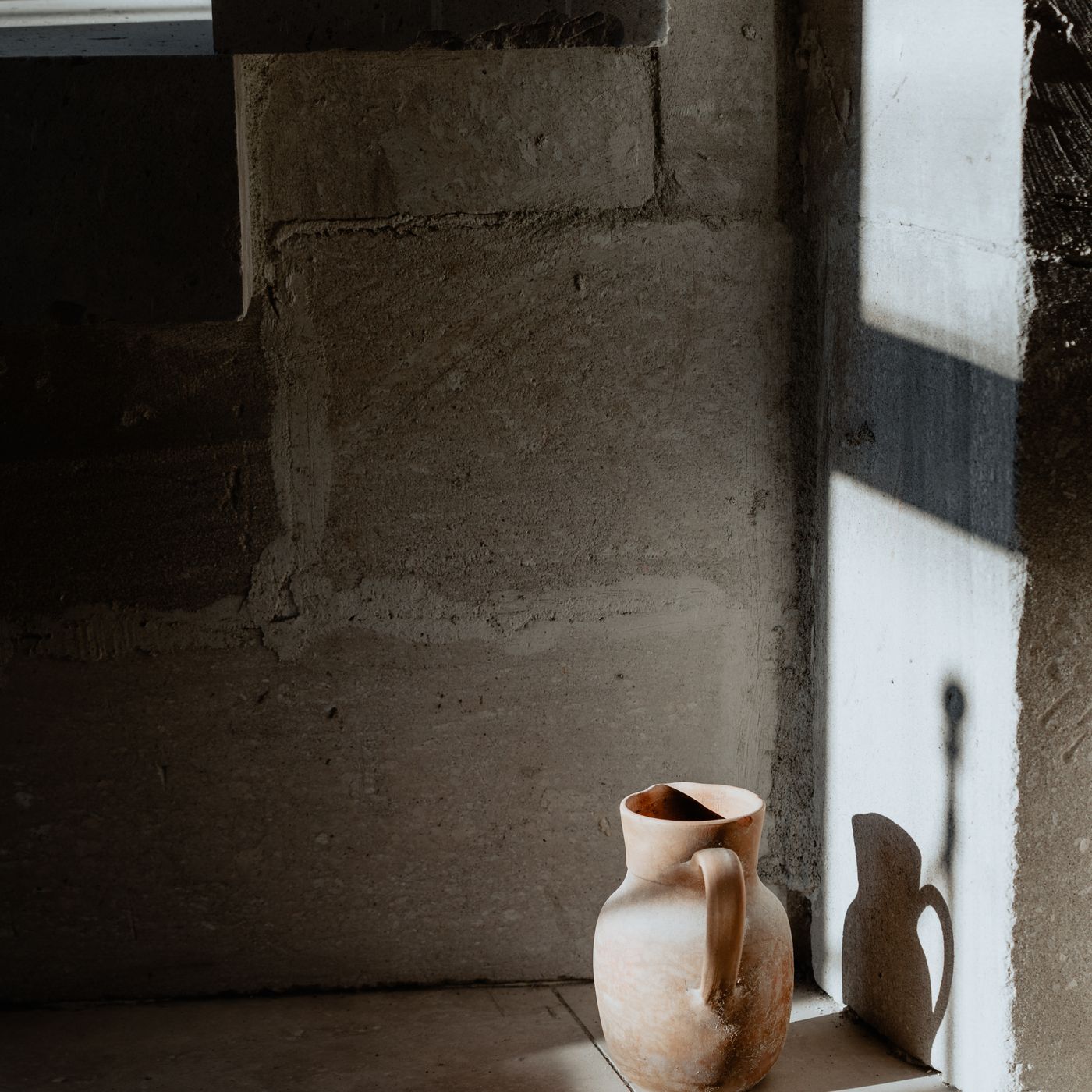 A rustic ceramic jug standing in sunlight by a stone wall, casting a long shadow.