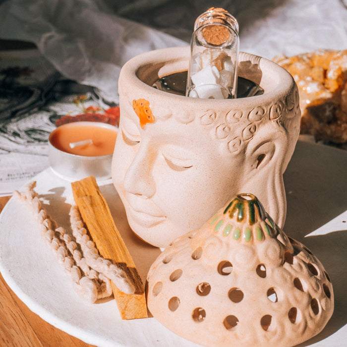 An incense holder shaped like a seated Buddha, shown with a candle and a few natural elements around it.