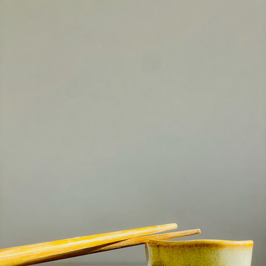 A simple ceramic bowl with chopsticks resting across it, set on a woven mat against a grey background.
