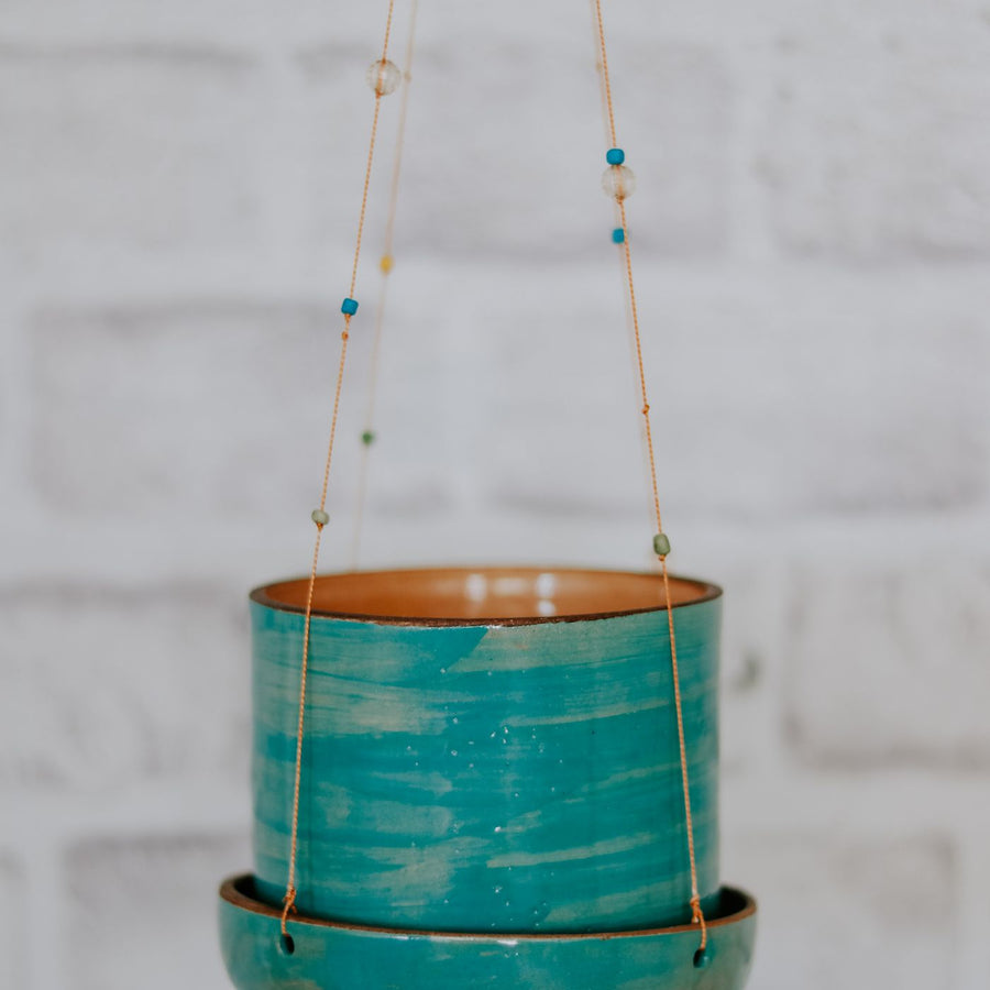 A teal ceramic hanging flower pot photographed close up against a brick wall.