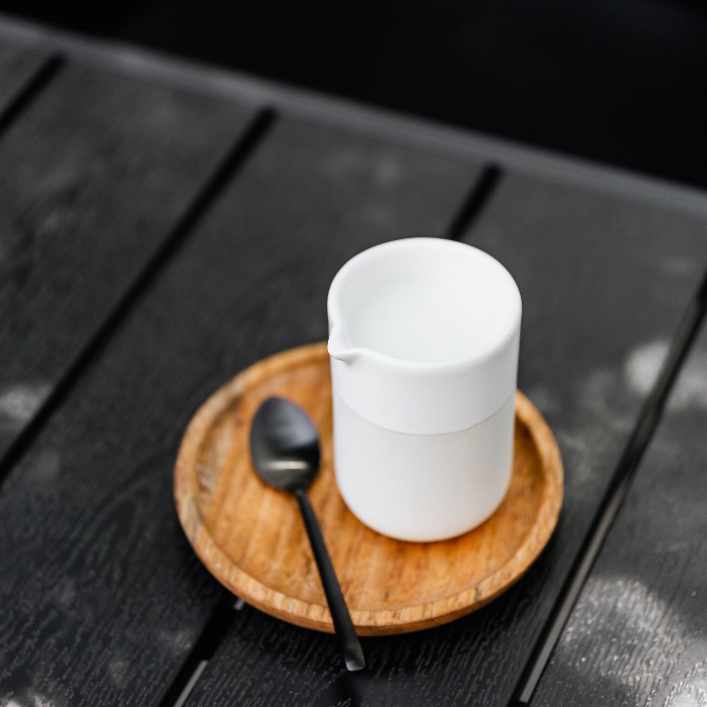 A ceramic milk jug seen close up with a spoon on a wooden tray, on a dark table.