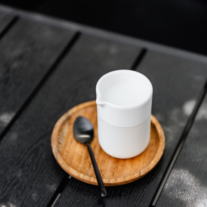 A ceramic milk jug seen close up with a spoon on a wooden tray, on a dark table.