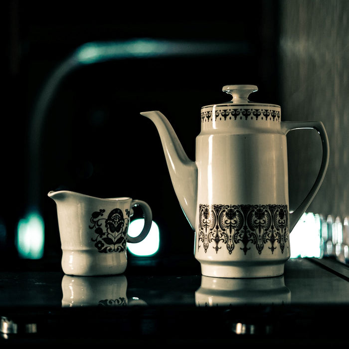 A ceramic teapot and a matching milk jug, both closely patterned, against a dim background.