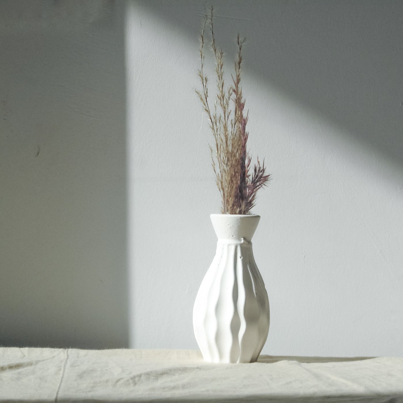 A plain white vase holding dried grass, shown against a grey background.