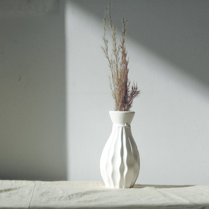 A plain white vase holding dried grass, shown against a grey background.