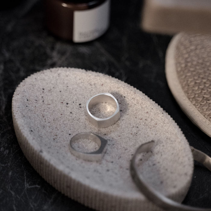A textured soap dish shown close up with minimalist silver jewellery laid on it.